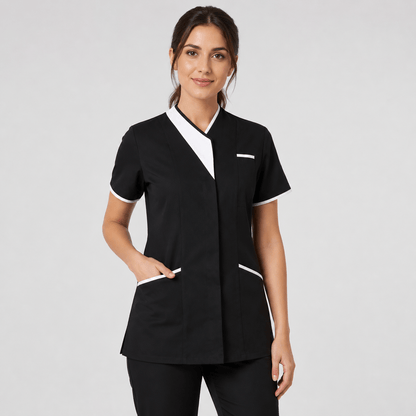 A woman modeling a black short-sleeve medical tunic with white contrast trim on the V-neck, sleeve cuffs, and pocket edges, paired with black scrub pants against a light gray studio background